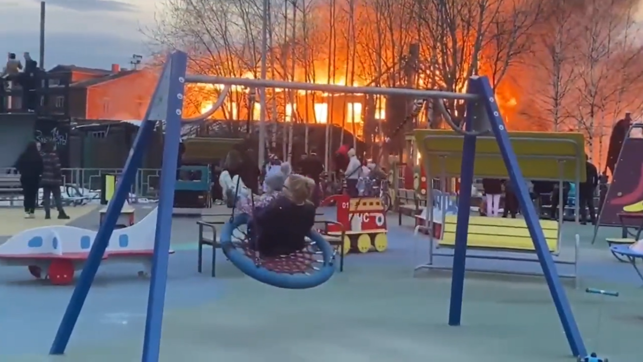 Russian woman casually swings with child as building burns nearby