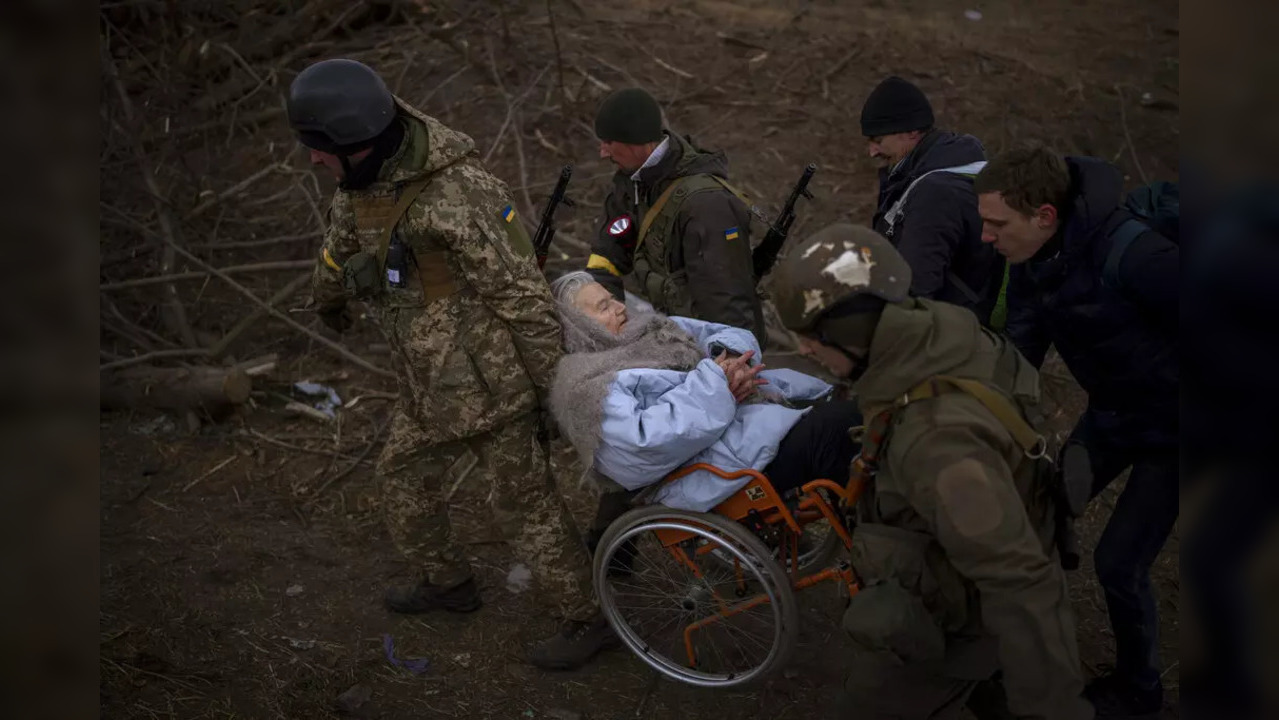 Ukrainian soldiers and militiamen carry a woman in a wheelchair as the artillery echoes nearby, while people flee Irpin on the outskirts of Kyiv