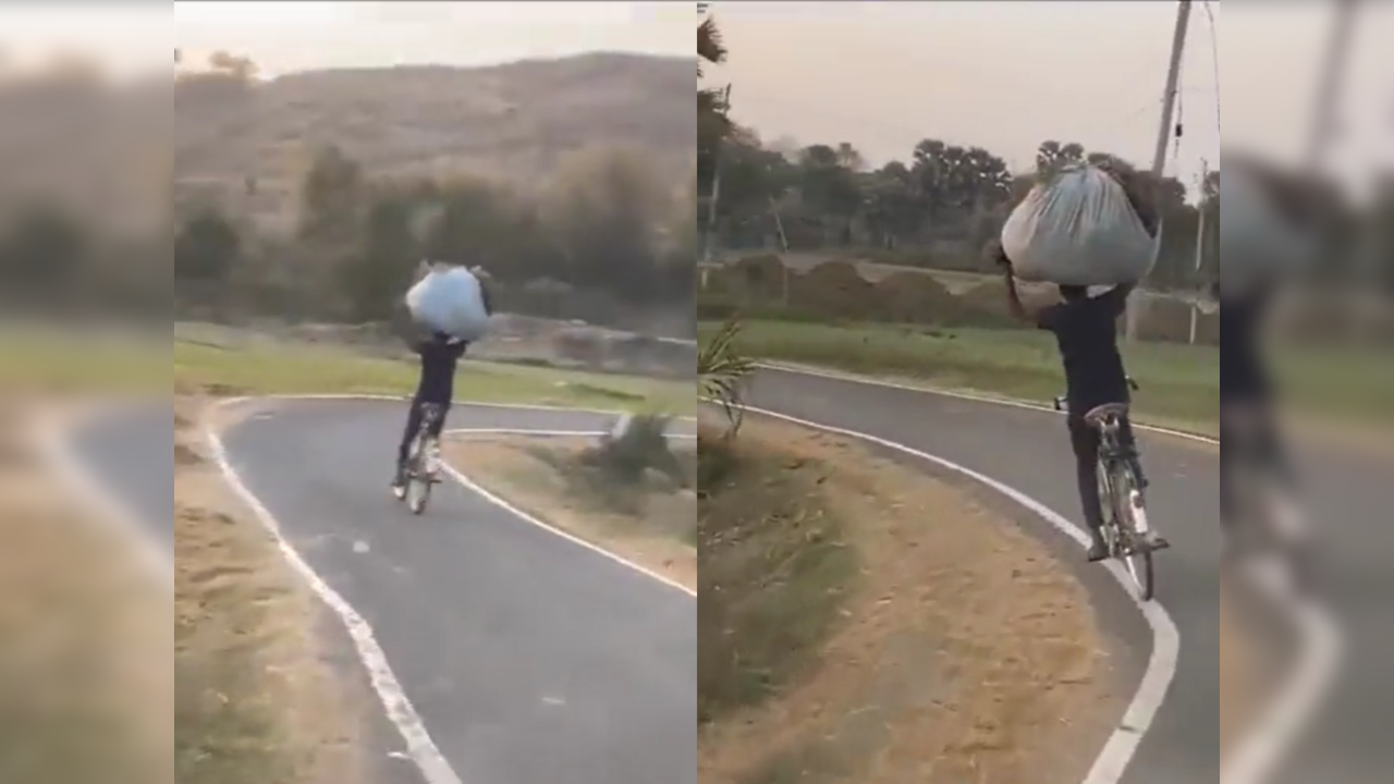 Man balances sack on head with both hands while cycling on narrow road